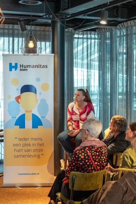 Ervaringsdeskundige vertelt haar verhaal op een kruk naast de Humanitas DMH-banner, fotografie bij de promotiefilm van Humanitas DMH