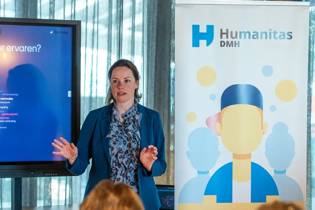 Spreker in blauwe blazer geeft een presentatie naast de Humanitas DMH-banner, fotografie bij de promotiefilm van Humanitas DMH
