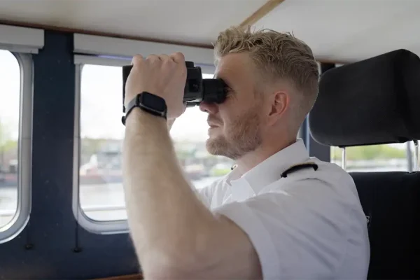 Havens en vaarwegen Zaanstad — schipper aan het werk Schipper in wit uniform kijkt door een verrekijker vanuit de stuurhut, beeld uit de wervingsfilm over werken bij havens en vaarwegen van gemeente Zaanstad