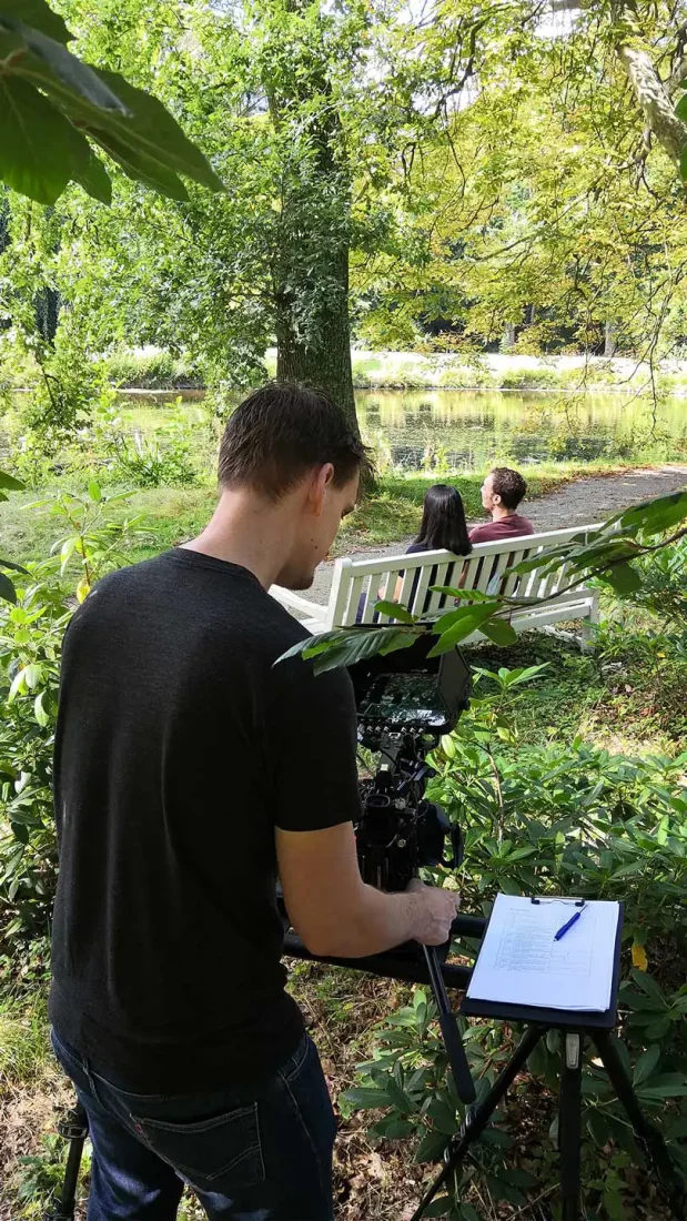 Moodfilm Hoge Vrijheid — opname in het park Cameraman van Mondha Media filmt twee mensen op een witte bank aan het water in het park voor de moodfilm Hoge Vrijheid