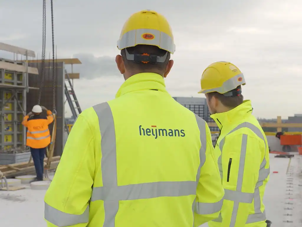 Twee medewerkers in geel veiligheidshesje op de bouwplaats terwijl een collega materiaal aanpakt, still uit de testimonial voor KIVI