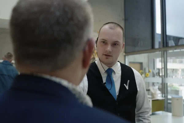 Testimonial VVNL — lid in gesprek met een bezoeker Medewerker in vest met das helpt een bezoeker in de museumhal, still uit de video van Mondha Media voor VVNL