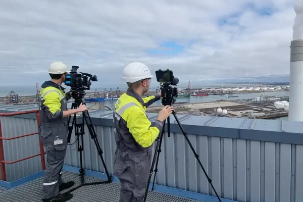 Twee cameramannen in veiligheidskleding en witte helmen filmen met statieven vanaf een hoog dak met uitzicht op een industriegebied en de haven voor de Uniper projectfilm.