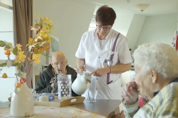 Samen aan tafel – zorgvideo De Waterpoort Verzorgende schenkt drinken in voor twee bewoners aan tafel in de woonkamer van De Waterpoort