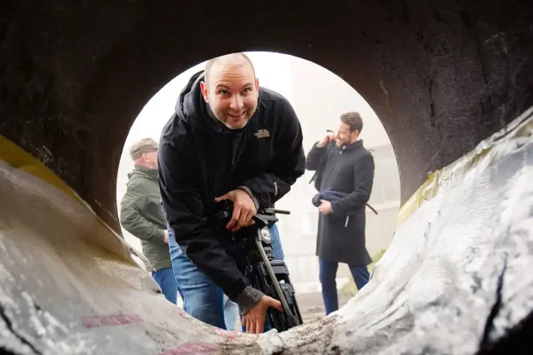 Creatief cameraperspectief bij B2B filmopdracht Een glimlachende Sebastiaan van Mondha Media kijkt door een grote ronde bouwbuis tijdens een B2B filmopdracht voor Vink Bouw Kings Amsterdam.