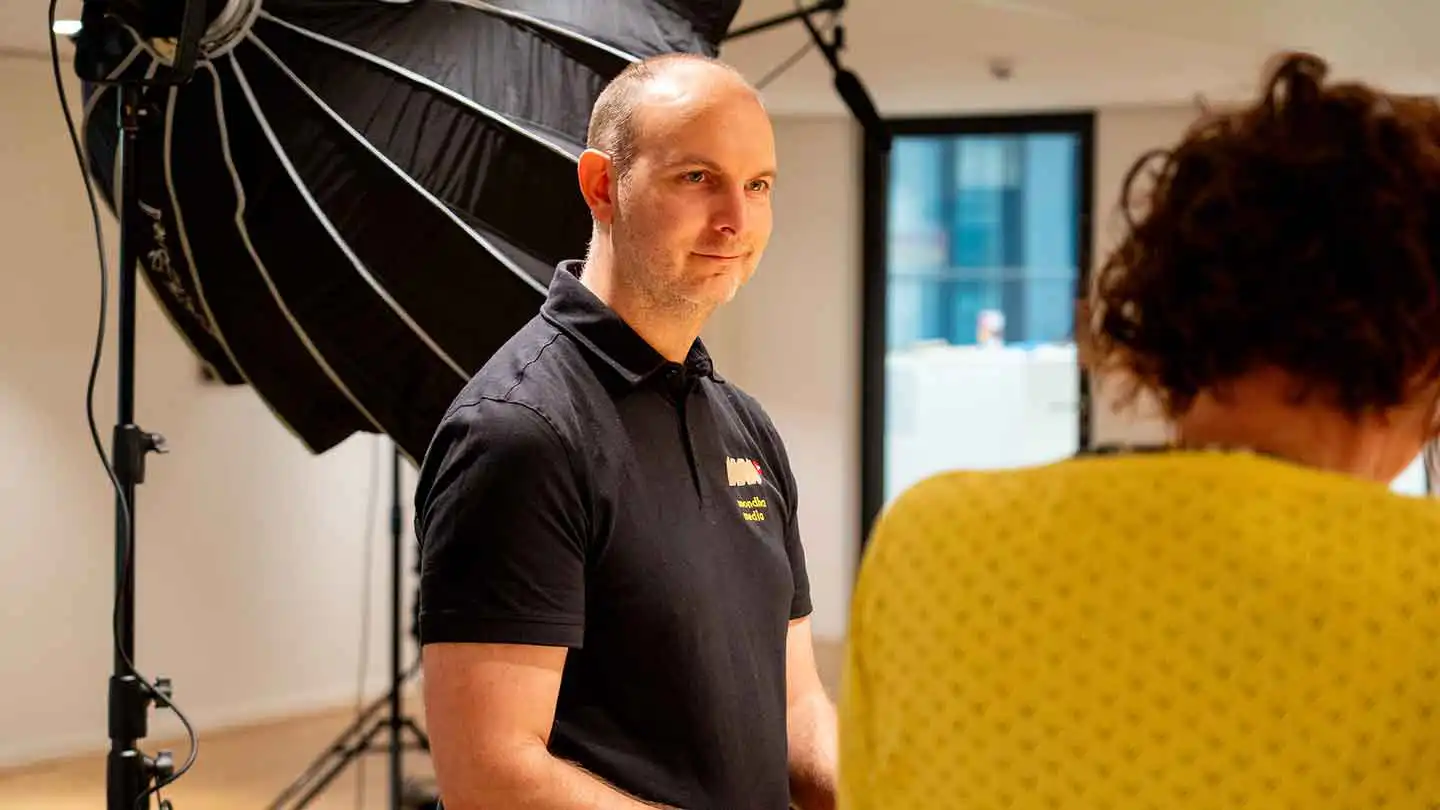 Sebastiaan van Mondha Media in een zwart poloshirt kijkt glimlachend naar een spreker op de filmset bij het ministerie van SZW, met een grote studiolamp op de achtergrond.