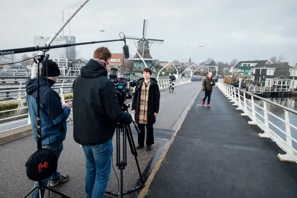 Filmcrew van Mondha Media filmt een medewerker van gemeente Zaanstad op een brug met een Zaanse molen op de achtergrond